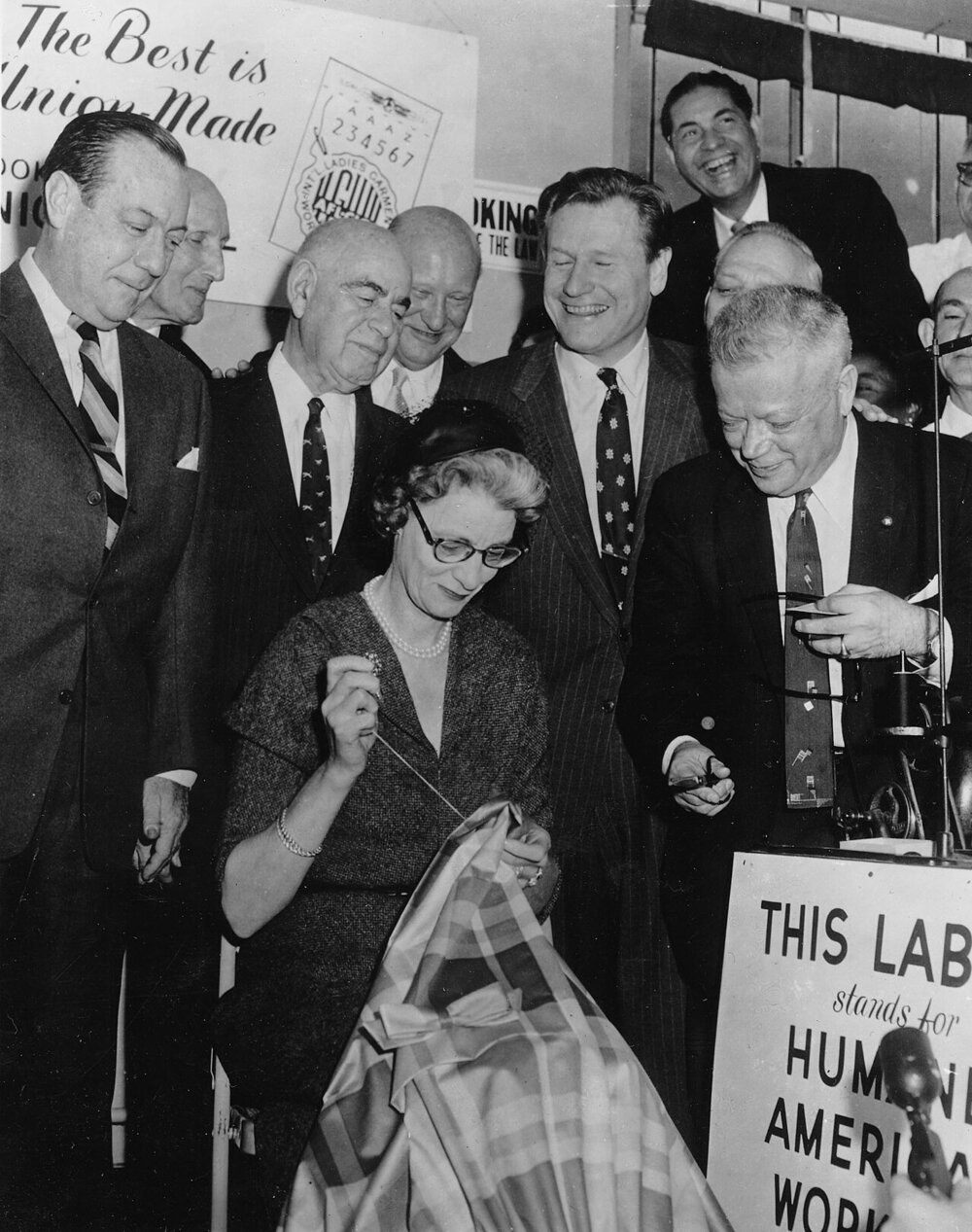 Black and white photo of Mary Rockefeller sewing an ILGWU union label into a garment beneath an enlarged label poster